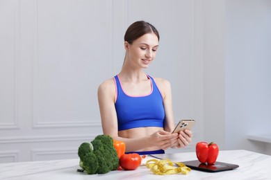 Photo of Woman using smartphone at white marble table with kitchen scale, vegetables and measuring tape indoors