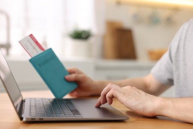 Photo of Man with passport and flight ticket using laptop at wooden table indoors, closeup