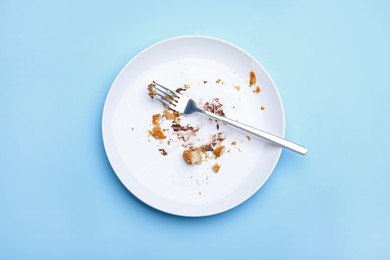 Photo of Dirty plate with crumbs, cream and fork on light blue background, top view