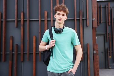 Photo of Teenage boy with headphones and backpack near building outdoors