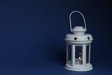 Photo of Beautiful white decorative lantern with burning candle on blue background, space for text