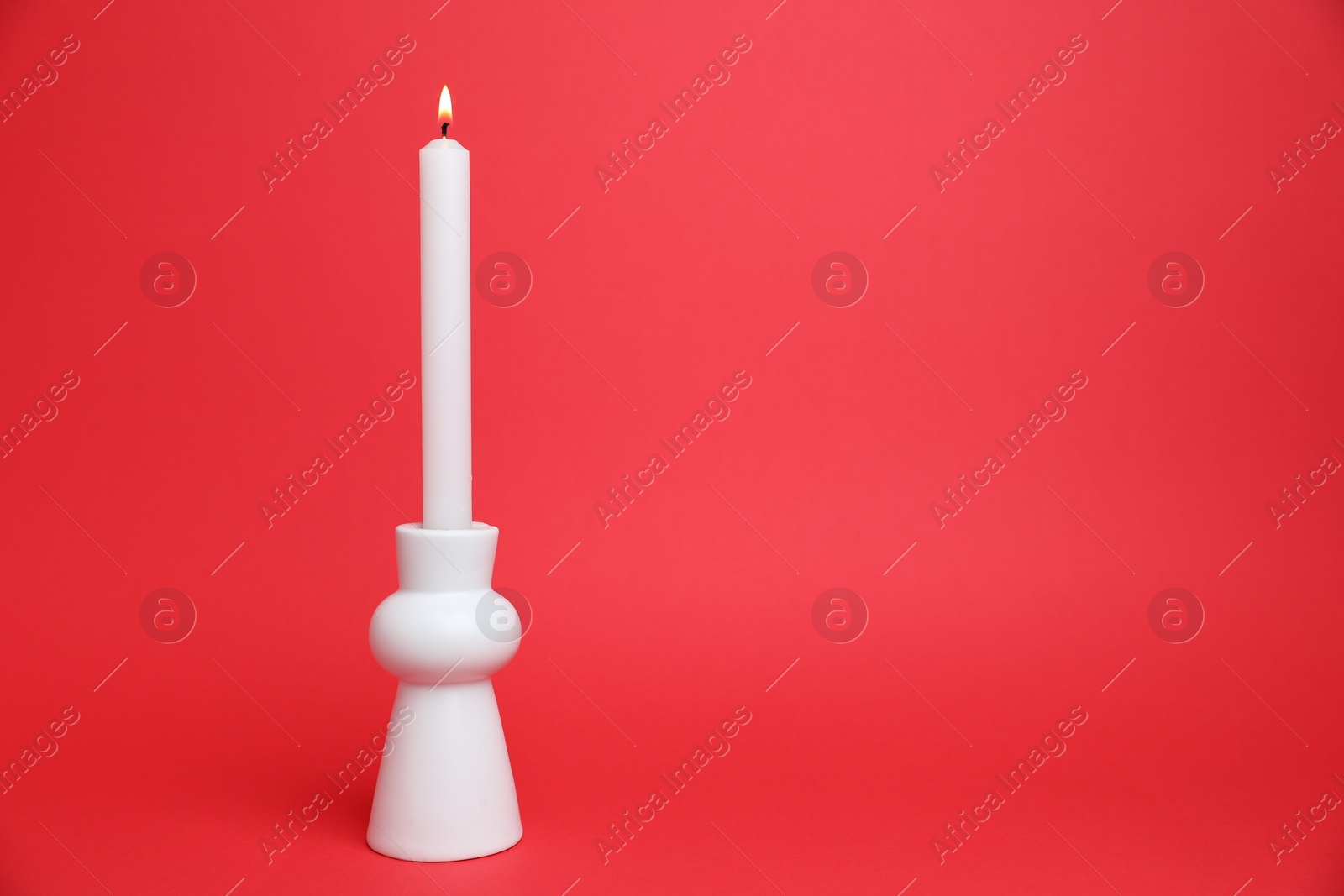 White candlestick with burning candle on red background, space for text Photo of White candlestick with burning candle on red background, space for text