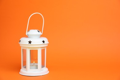 Photo of Beautiful white decorative lantern with burning candle on orange background, space for text