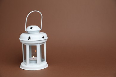 Photo of Beautiful white decorative lantern with burning candle on brown background, space for text