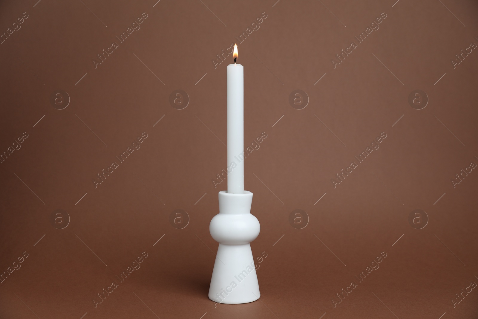 White candlestick with burning candle on brown background Photo of White candlestick with burning candle on brown background