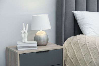 Photo of Lamp, white figure in shape of hand and books on bedside table near bed indoors