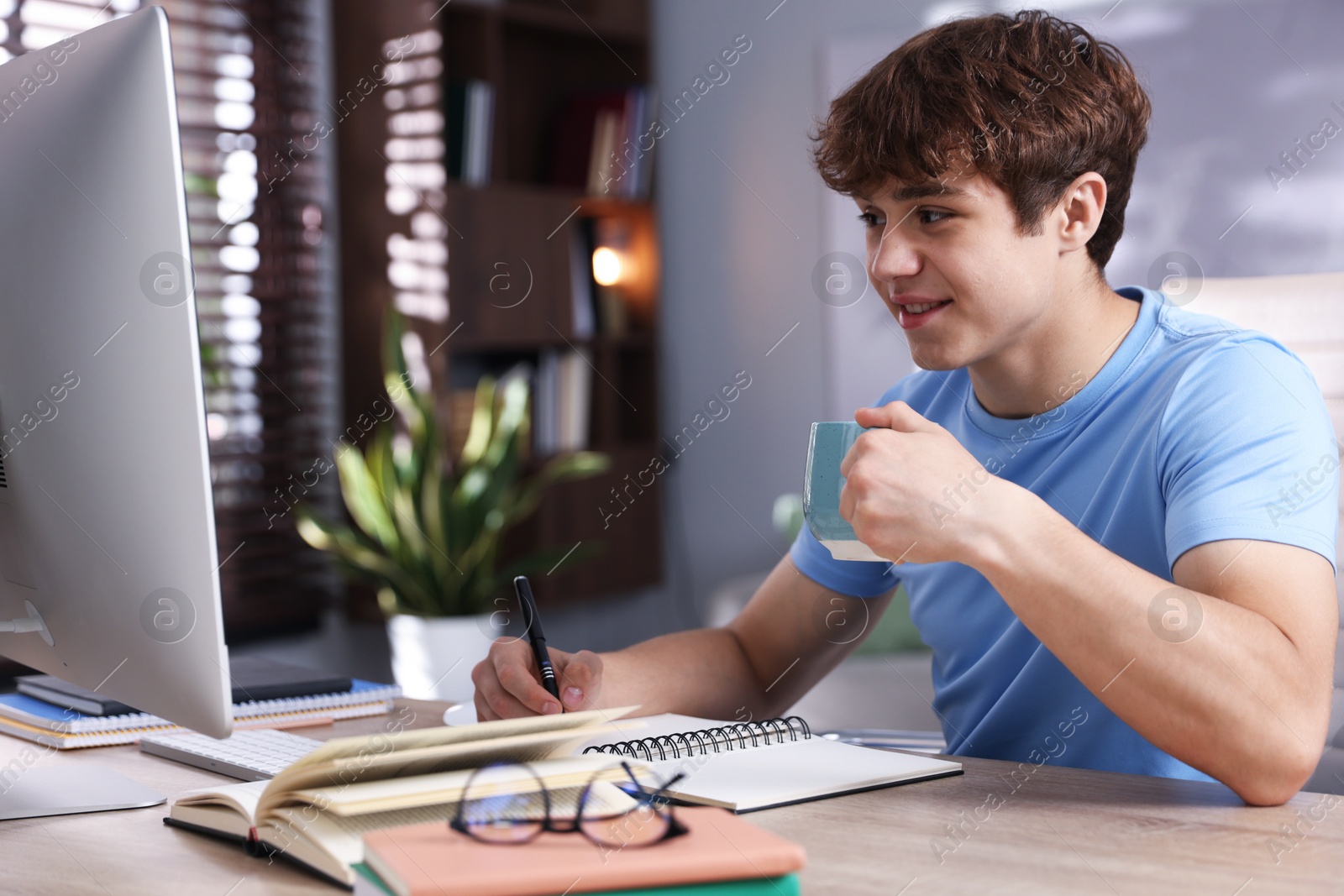 Teenage student with cup of drink taking notes while studying at table indoors Photo of Teenage student with cup of drink taking notes while studying at table indoors