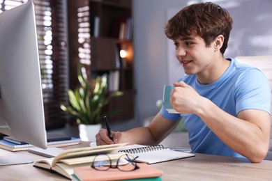 Photo of Teenage student with cup of drink taking notes while studying at table indoors