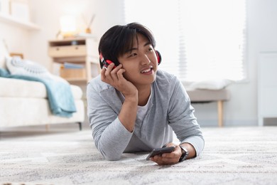 Photo of Smiling Asian man with headphones and smartphone on floor at home
