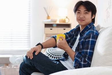 Photo of Handsome Asian man with cup of hot drink on sofa at home