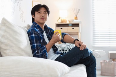 Photo of Handsome Asian man with cup of hot drink on sofa at home