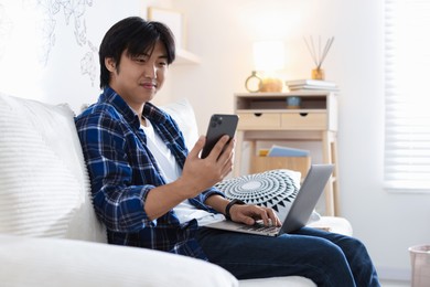 Photo of Handsome Asian man with smartphone and laptop on sofa at home