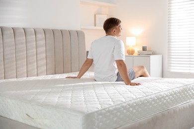 Photo of Young man sitting on bed with soft mattress at home, back view
