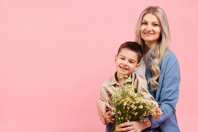 Photo of Little boy and his smiling mother with bouquet of beautiful chamomile flowers on pink background. Space for text