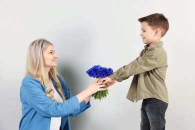 Photo of Little boy presenting bouquet of beautiful cornflowers to his smiling mother on light grey background