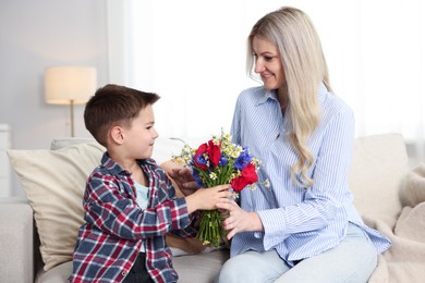 Photo of Little boy presenting bouquet of beautiful flowers to his smiling mother on sofa at home