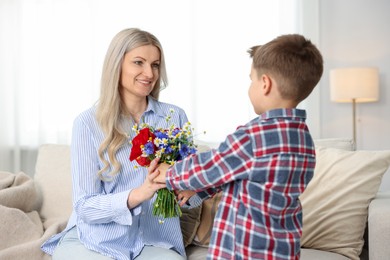 Photo of Little boy presenting bouquet of beautiful flowers to his smiling mother on sofa at home