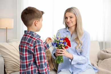 Photo of Little boy presenting bouquet of beautiful flowers to his smiling mother on sofa at home