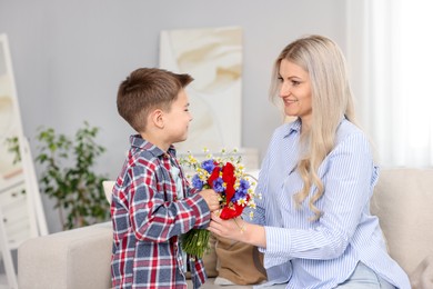Photo of Little boy presenting bouquet of beautiful flowers to his smiling mother on sofa at home