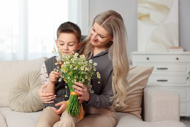 Photo of Little boy and his smiling mother with bouquet of beautiful chamomile flowers on sofa at home