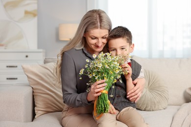 Photo of Little boy and his mother with bouquet of beautiful chamomile flowers on sofa at home
