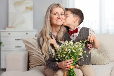 Photo of Little boy kissing his smiling mother with bouquet of beautiful chamomile flowers on sofa at home