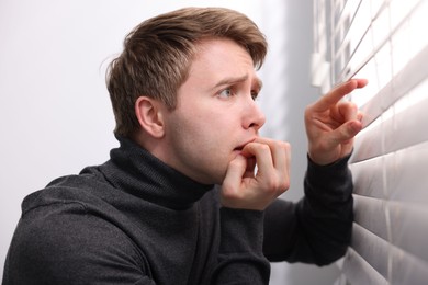 Photo of Paranoia. Worried man looking through window blinds indoors