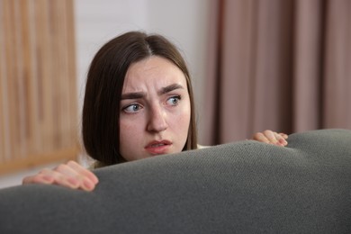 Photo of Paranoia. Worried woman hiding behind sofa indoors