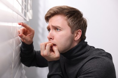 Photo of Paranoia. Worried man looking through window blinds indoors