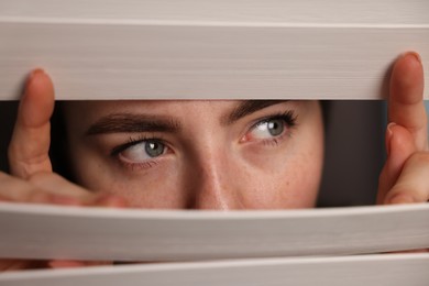 Photo of Paranoia. Worried woman looking through window blinds indoors, closeup