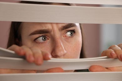 Photo of Paranoia. Worried woman looking through window blinds indoors, closeup