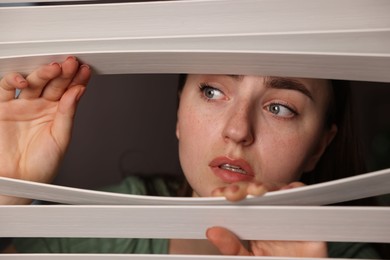 Photo of Paranoia. Worried woman looking through window blinds indoors