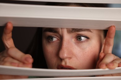 Photo of Paranoia. Worried woman looking through window blinds indoors