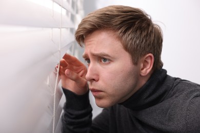 Photo of Paranoia. Worried man looking through window blinds indoors