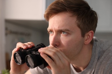 Photo of Paranoia. Worried man looking through binoculars in kitchen