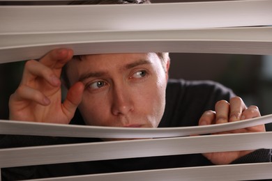 Photo of Paranoia. Worried man looking through window blinds indoors