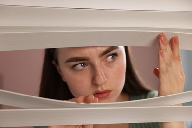 Photo of Paranoia. Worried woman looking through window blinds indoors