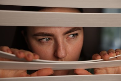 Photo of Paranoia. Worried woman looking through window blinds indoors, closeup