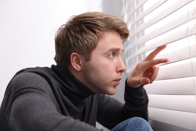 Photo of Paranoia. Worried man looking through window blinds indoors