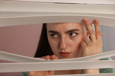 Photo of Paranoia. Worried woman looking through window blinds indoors