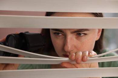 Photo of Paranoia. Worried woman with binoculars looking through window blinds indoors