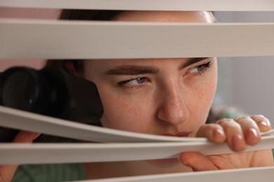 Photo of Paranoia. Worried woman with binoculars looking through window blinds indoors