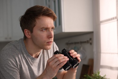 Photo of Paranoia. Worried man with binoculars in kitchen, space for text