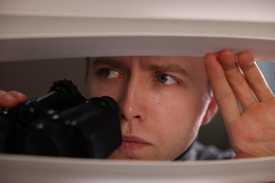 Photo of Paranoia. Worried man with binoculars looking through window blinds indoors