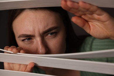 Photo of Paranoia. Worried woman looking through window blinds indoors