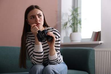 Photo of Paranoia. Worried woman with binoculars on sofa at home