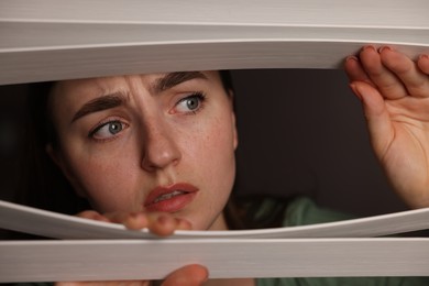 Photo of Paranoia. Worried woman looking through window blinds indoors