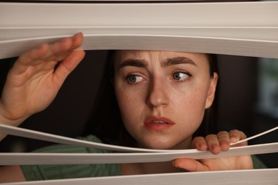 Photo of Paranoia. Worried woman looking through window blinds indoors