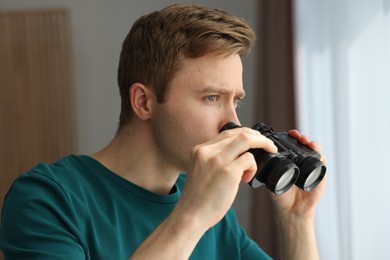 Photo of Paranoia. Worried man with binoculars near window indoors
