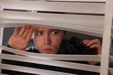 Photo of Paranoia. Worried man looking through window blinds indoors